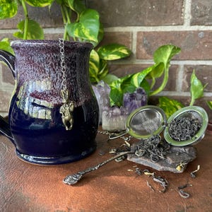 May include: A handmade ceramic mug with a dark blue base and a purple and brown rim. A tea infuser with loose tea sits next to the mug. A small decorative spoon and a charm hanging from the mug complete the tea set.
