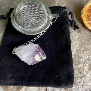 May include: A silver tea infuser with a chain and a purple amethyst crystal. The infuser is sitting on a black velvet pouch.