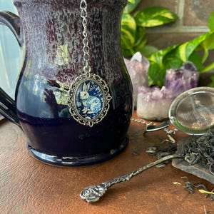 May include: A dark blue and purple ceramic mug with a silver tea infuser featuring a white rabbit design. A decorative spoon and tea strainer are also visible, suggesting a tea-related theme.