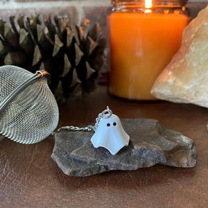 May include: A silver mesh tea infuser and a small white ghost charm with black dot eyes sit on a dark stone. A lit orange candle and a pine cone are in the background. The charm is attached to a silver chain.