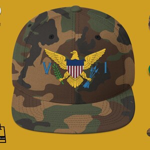 Puede incluir: Gorra de béisbol de camuflaje con el emblema de las Islas Vírgenes. El emblema incluye un águila dorada, un escudo con rayas rojas y blancas, y las letras "V" e "I" en azul. La gorra tiene una visera verde.