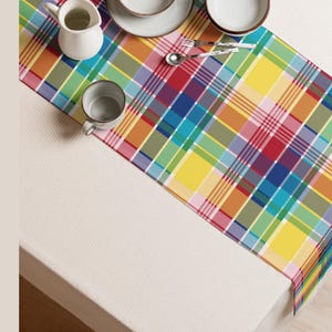 May include: A colorful plaid table runner in a rainbow of hues, including red, yellow, green, and blue. The runner is placed on a white tablecloth, with tableware and a small pitcher visible.