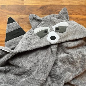 May include: A gray hooded towel with a raccoon face design. The towel features a hood with a raccoon face, including eyes, a nose, and ears. The tail is black and gray striped. The towel is made of a soft, absorbent material.