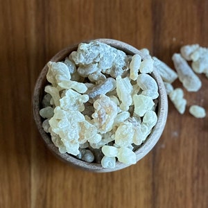 Puede incluir: Un cuenco de madera lleno de trozos de resina de color amarillo pálido y blanquecino. Algunos trozos están esparcidos fuera del cuenco sobre una superficie de madera. La resina tiene un aspecto ligeramente translúcido.