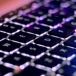 Può includere: Primo piano di una tastiera di laptop nera con tasti bianchi e una luce blu che si riflette sui tasti.