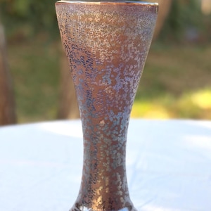 May include: A tall, flared ceramic vase with a metallic, iridescent brown and blue crystalline glaze. The vase features a wide, open rim that tapers down to a narrow neck and widens at the base, which is supported by three small, decorative feet.