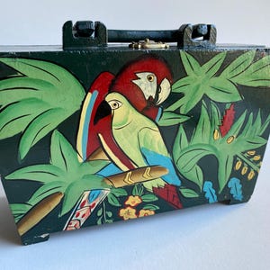 May include: A wooden box with a hinged lid and a handle. The box is painted with a tropical scene featuring two parrots, green leaves, and flowers. The parrots are red, green, and blue.