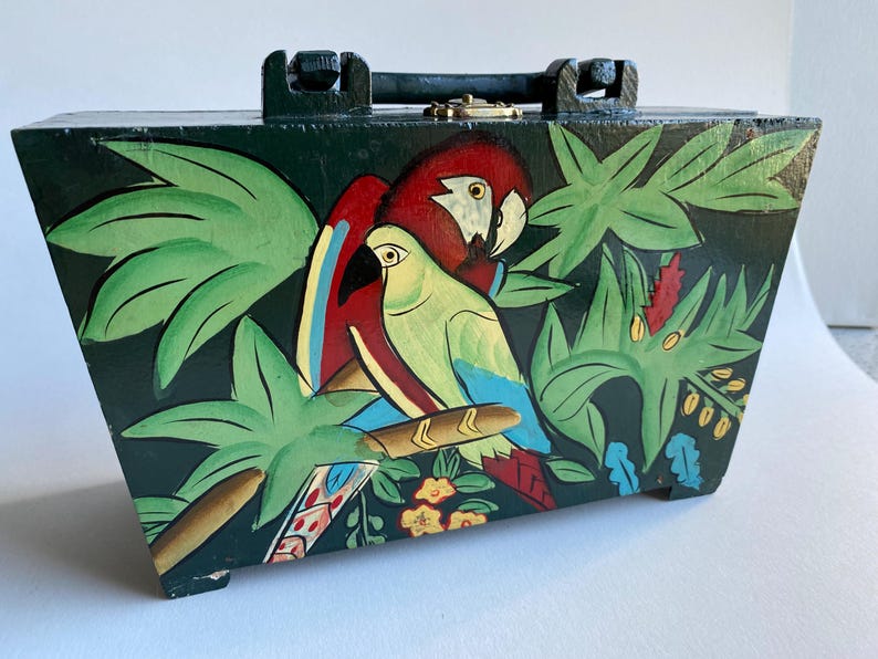 May include: A wooden box with a hinged lid and a handle. The box is painted with a tropical scene featuring two parrots, green leaves, and flowers. The parrots are red, green, and blue.