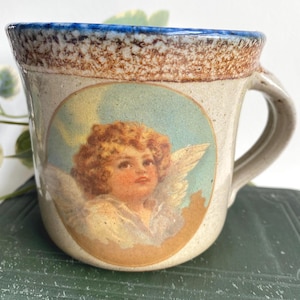 Puede incluir: Una taza de cerámica con base beige claro, borde moteado marrón y borde superior azul. La taza presenta una ilustración vintage de un querubín con cabello dorado, alas y una expresión serena. El asa es de color beige.