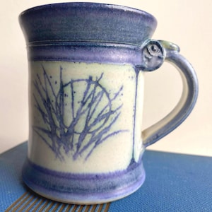 Puede incluir: Taza de cerámica hecha a mano en azul y blanco. La taza tiene un cuerpo cilíndrico, un asa y un borde acampanado. El cuerpo está decorado con un diseño abstracto azul. La taza mide aproximadamente 10 cm de alto.
