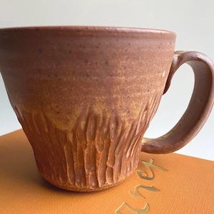 Puede incluir: Una taza de cerámica color terracota moteada con un borde ancho y acampanado y un asa curva. La parte inferior de la taza presenta un patrón vertical tallado y texturizado que contrasta con la sección superior más lisa y el asa.