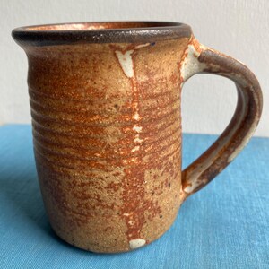 Peut inclure: Une tasse en céramique avec un vernis moucheté beige et brun, présentant un corps cylindrique avec des textures striées horizontales, un bord brun foncé et une anse incurvée. La surface affiche une finition terreuse avec des accents blancs.