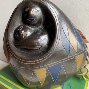 May include: A ceramic sculpture of two figures nestled together inside a larger, egg-shaped form. The sculpture is painted in shades of black, blue, gold, and white, with geometric patterns on the exterior.