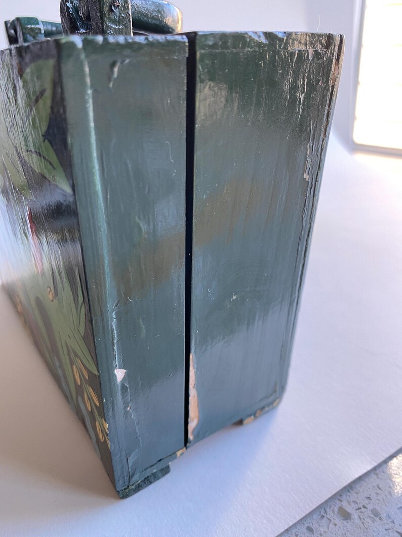 May include: A green wooden box with a hinged lid and painted floral details. The box is closed and the lid is slightly ajar.