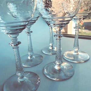 Op de afbeelding: Een verzameling van vijf heldere, geëtste kristallen champagneglazen. De glazen hebben een delicaat bloemmotief op de kelk en een diamantpatroon op de steel. De bases zijn rond en helder. De glazen zijn gerangschikt op een licht oppervlak.