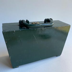 May include: A dark green wooden box with a brass latch and a black handle. The box has a rectangular shape and is sitting on a white surface.