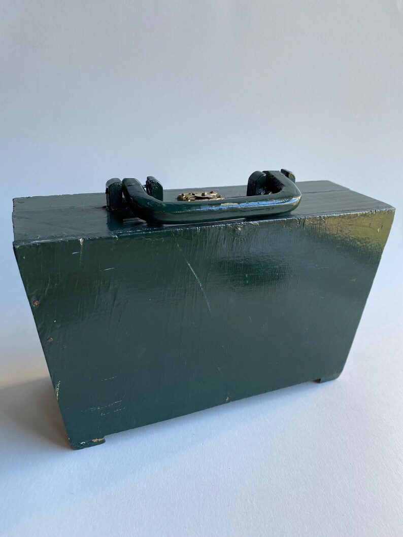May include: A dark green wooden box with a brass latch and a black handle. The box has a rectangular shape and is sitting on a white surface.