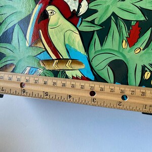 May include: A wooden box with a painted design of tropical leaves and parrots. The box is approximately 30 inches long and has a black base.