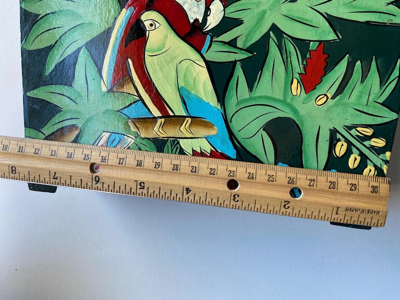 May include: A wooden box with a painted design of tropical leaves and parrots. The box is approximately 30 inches long and has a black base.