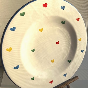 May include: A round ceramic plate with a white base and a thin blue rim. The surface of the plate is decorated with a scattered pattern of small, hand-painted hearts in primary colors including red, blue, yellow, and green across the entire face.