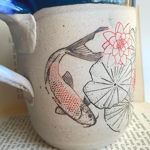 May include: A ceramic mug with a white speckled finish, featuring a koi fish and lotus flower design. The fish is red and the flowers are red and white. The rim is dark blue, and the handle is white.