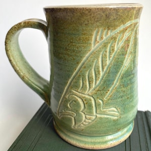 May include: Green ceramic mug with a carved fish design. The mug has a handle and a wide base. The fish design is carved into the side of the mug, and the mug has a speckled finish. The rim of the mug is a darker brown colour.