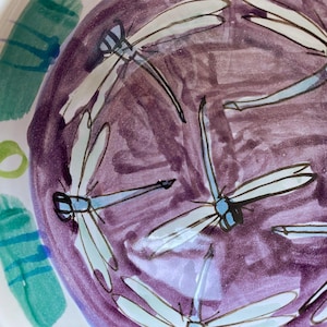 May include: A ceramic bowl with a purple interior and a white exterior. The bowl is decorated with hand-painted dragonflies in white, light blue, and black. Green and blue abstract shapes are painted on the rim.