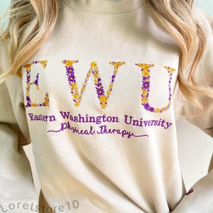 Könnte beinhalten: Ein beiger Pullover mit den Buchstaben "EWU" in lila und gelber Blumenstickerei. Der Text "Eastern Washington University Physical Therapy" ist unter den Buchstaben gestickt.