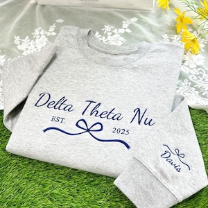 May include: Gray sweatshirt with a navy blue embroidered design that says "Delta Theta Nu EST. 2025" with a bow. The right sleeve has a navy blue embroidered bow with the word "Davis" underneath.