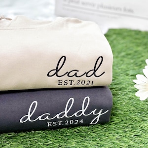 May include: Two stacked sweatshirts, one tan and one dark gray. The tan sweatshirt has the word "dad" embroidered in cursive with "EST.2021" below. The dark gray sweatshirt has "daddy" embroidered in cursive with "EST.2024" below.