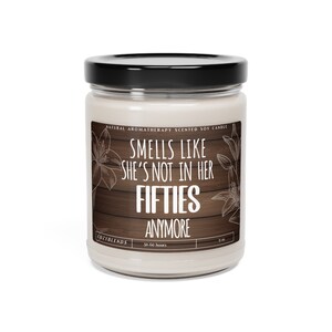 May include: A white soy candle with a black lid and a wooden background. The candle has the text "Smells like she's not in her fifties anymore" on the label. The candle is 9 oz and has a burn time of 50-60 hours.