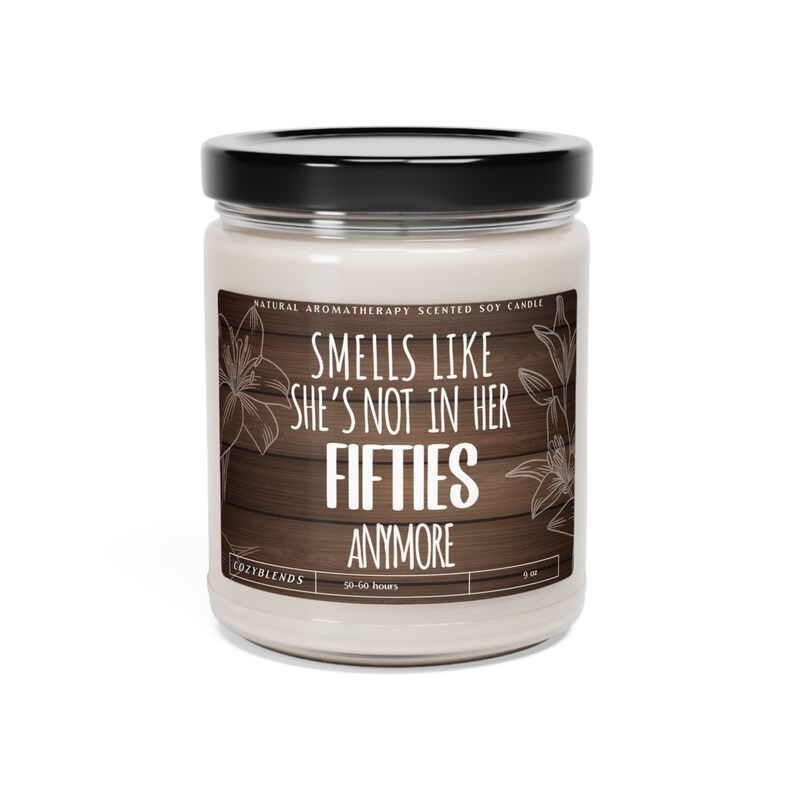 May include: A white soy candle with a black lid and a wooden background. The candle has the text "Smells like she's not in her fifties anymore" on the label. The candle is 9 oz and has a burn time of 50-60 hours.