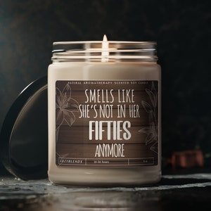 May include: A beige soy candle with a wooden label that reads "Smells like she's not in her fifties anymore". The candle is in a clear glass jar with a black lid. The label also says "Natural Aromatherapy Scented Soy Candle" and "Cozyblends".