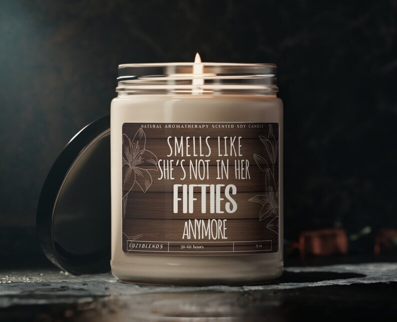 May include: A beige soy candle with a wooden label that reads "Smells like she's not in her fifties anymore". The candle is in a clear glass jar with a black lid. The label also says "Natural Aromatherapy Scented Soy Candle" and "Cozyblends".