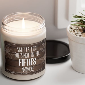 May include: A white soy candle with a brown label that reads "Smells like she's not in her fifties anymore". The candle is lit and has a natural aromatherapy scent.