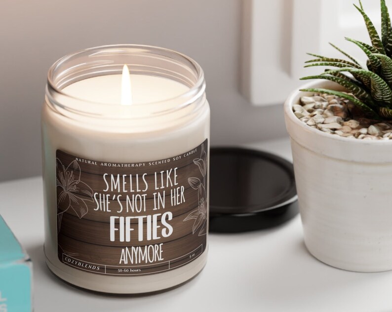 May include: A white soy candle with a brown label that reads "Smells like she's not in her fifties anymore". The candle is lit and has a natural aromatherapy scent.