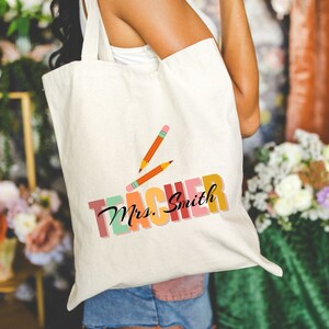 Pu&ograve; includere: Una borsa di tela bianca con il testo "Teacher" in lettere colorate e le parole "Mrs. Smith" in corsivo nero. Due matite gialle sono disegnate sopra il testo.
