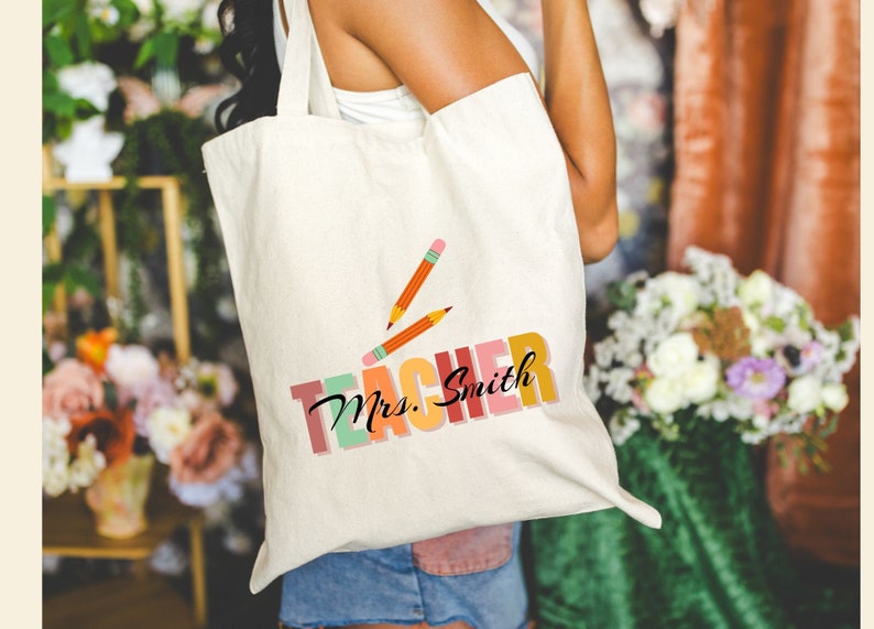 Pu&ograve; includere: Una borsa di tela bianca con il testo "Teacher" in lettere colorate e le parole "Mrs. Smith" in corsivo nero. Due matite gialle sono disegnate sopra il testo.