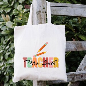 Pu&ograve; includere: Una borsa di tela bianca con il testo "Teacher" in colori arcobaleno e il nome "Mrs. Smith" in nero. Due matite arancioni sono disegnate sopra il testo.