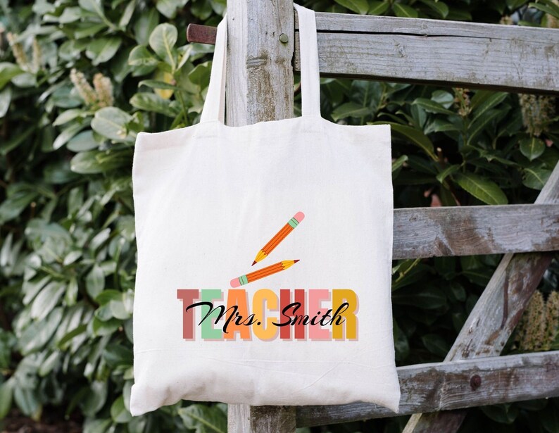 Pu&ograve; includere: Una borsa di tela bianca con il testo "Teacher" in colori arcobaleno e il nome "Mrs. Smith" in nero. Due matite arancioni sono disegnate sopra il testo.