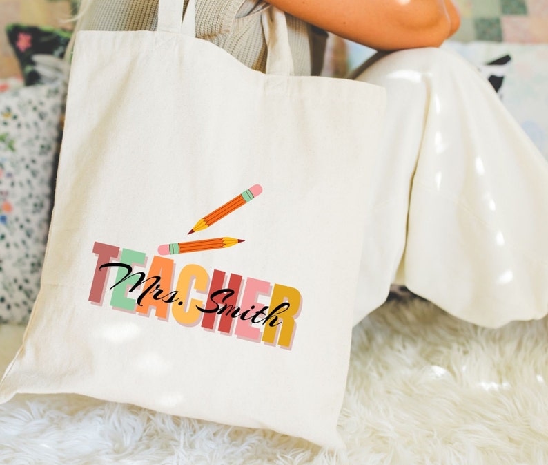 Pu&ograve; includere: Una borsa di tela bianca con il testo "TEACHER Mrs. Smith" in lettere colorate. Due matite sono disegnate sulla borsa.
