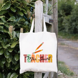 Pu&ograve; includere: Una borsa di tela bianca con un design "Teacher" colorato e il nome "Mrs. Smith" stampato su di essa. Due matite arancioni sono incrociate sulla parola "Teacher".