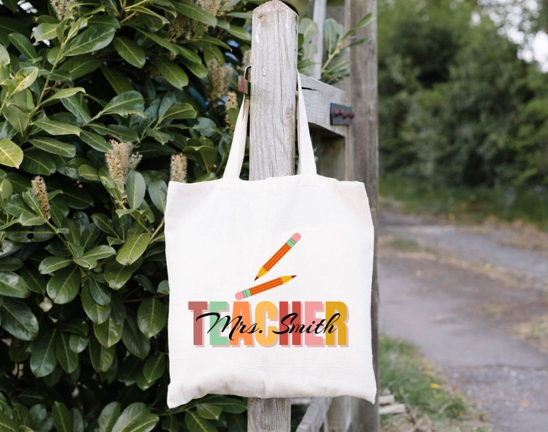 Pu&ograve; includere: Una borsa di tela bianca con un design "Teacher" colorato e il nome "Mrs. Smith" stampato su di essa. Due matite arancioni sono incrociate sulla parola "Teacher".