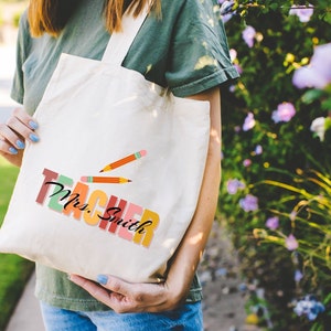Pu&ograve; includere: Una borsa tote in tela bianca con le parole "Teacher Mrs. Smith" in lettere maiuscole colorate e due matite gialle disegnate sulla borsa.