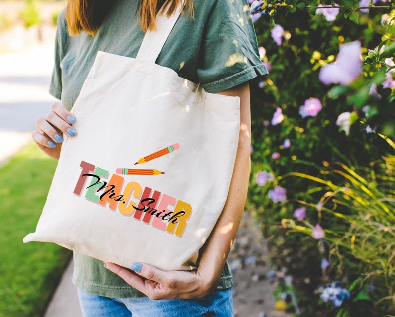 Pu&ograve; includere: Una borsa tote in tela bianca con le parole "Teacher Mrs. Smith" in lettere maiuscole colorate e due matite gialle disegnate sulla borsa.