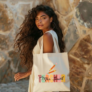 Pu&ograve; includere: Una borsa tote beige con un design "Teacher" colorato e il nome "Mrs. Smith" in corsivo. La borsa presenta due matite arancioni con gomme rosa.