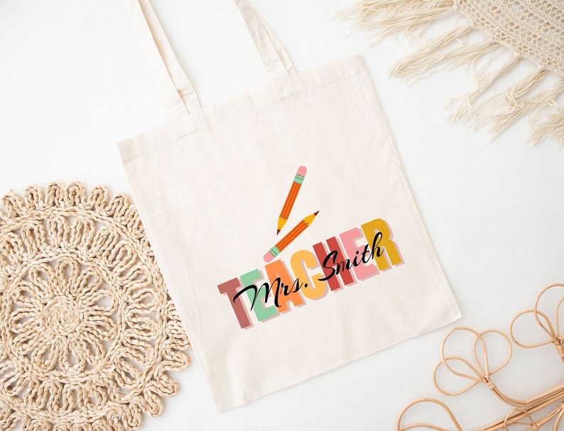 Pu&ograve; includere: Una borsa di tela bianca con il testo "Teacher" in colori arcobaleno e il nome "Mrs. Smith" in nero. Due matite sono mostrate sopra il testo.