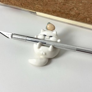 Puede incluir: Una figura de gato de cerámica blanca sosteniendo un cuchillo X-Acto plateado. El gato tiene una pequeña fresa beige en la cabeza. El cuchillo es plateado con el texto "X-ACTO 11" impreso en la hoja. La figura del gato está sobre una superficie blanca.