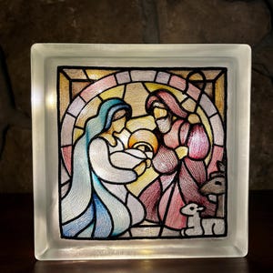 Puede incluir: Un bloque de vidrio con un diseño de vitral que representa la escena de la natividad. María, José y el niño Jesús están rodeados por un halo de luz. La escena está enmarcada en negro y el fondo es rosa claro y amarillo.