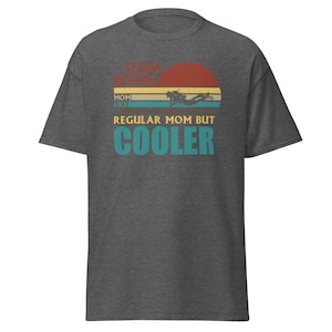 以下が含まれることがあります： 「Scuba Diving Mom Like Regular Mom But Cooler」とヴィンテージスタイルのグラフィックがプリントされたダークグレーのTシャツ。グラフィックには、夕焼けのシーンでスキンダイビングをしているダイバーが描かれている。